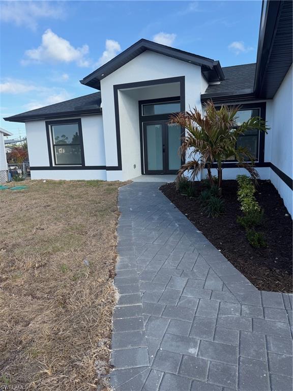 View of exterior entry with stucco siding, roof with shingles, and a lawn