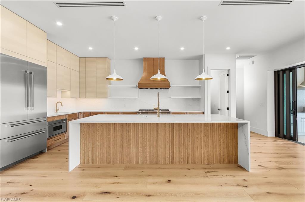Kitchen with stainless steel appliances, a kitchen island with sink, light wood finish cabinetry, and light stone countertops
