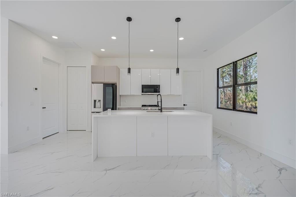 Kitchen featuring decorative light fixtures, an island with sink, stainless steel fridge, light stone counters, and white cabinets