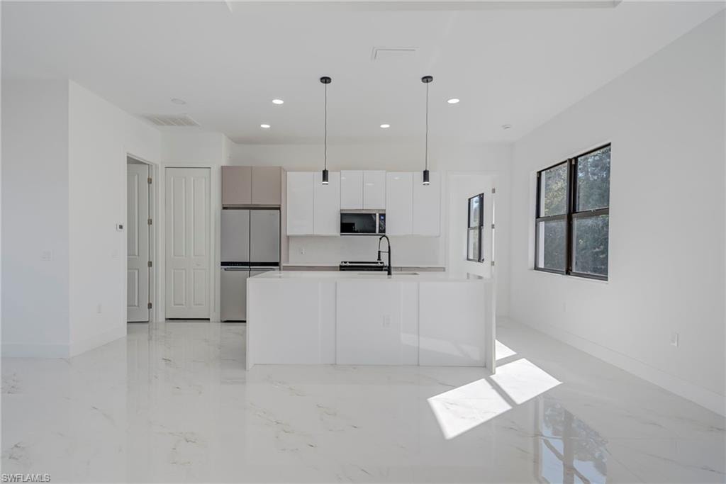 Kitchen featuring stainless steel appliances, light marble finish floors, a center island with sink, hanging light fixtures, and white cabinets