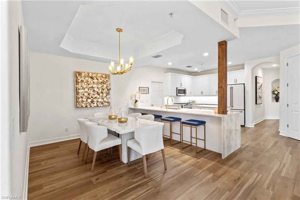 Dining room with arched walkways, light wood-type flooring, ornamental molding, a raised ceiling, and suspended lighting