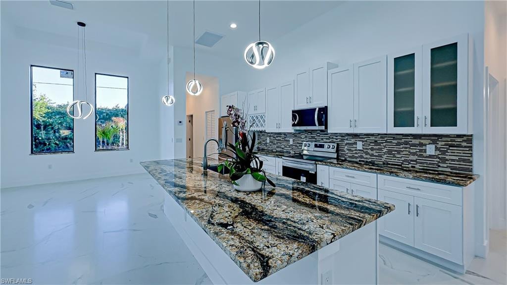 Kitchen featuring dark stone countertops, a kitchen island with sink, white cabinets, glass insert cabinets, and stainless steel appliances