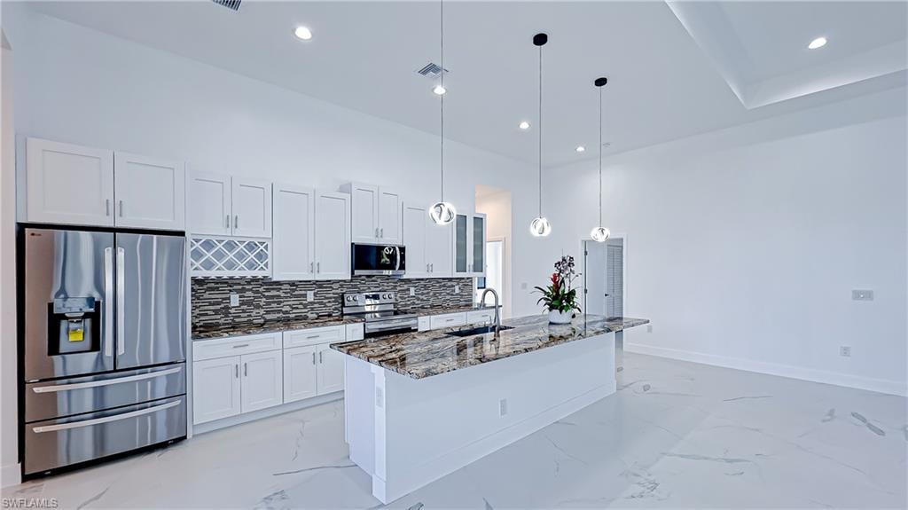 Kitchen with stainless steel appliances, dark stone countertops, hanging light fixtures, white cabinets, and a center island with sink