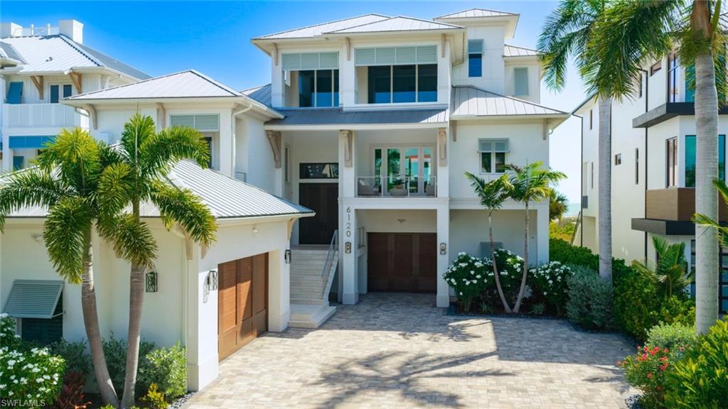 Multi-level coastal residence featuring a paver driveway, multiple garage doors with wood-finish panels, a second-story balcony, mature palm trees, and lush landscaping - Virtually Edited Image