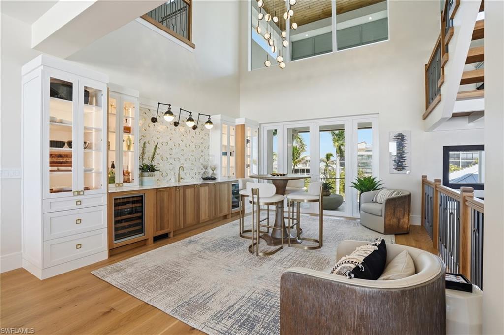 Indoor wet bar featuring glass fronted cabinets, light wood-style flooring, wine cooler, dual tone cabinetry, and a high ceiling - Virtually Edited Image