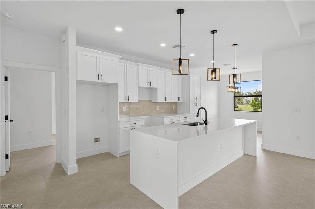 Kitchen featuring decorative light fixtures, light stone counters, an island with sink, decorative backsplash, and white cabinetry