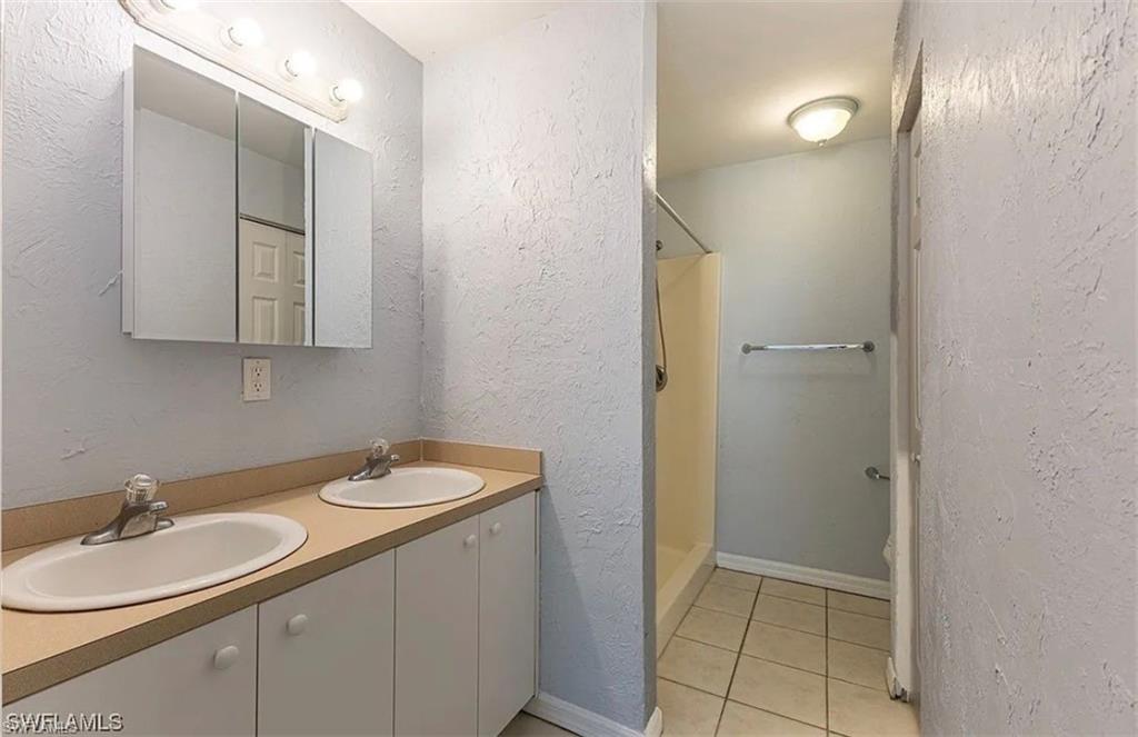 Full bathroom featuring a textured wall, double vanity, light tile patterned floors, and a stall shower