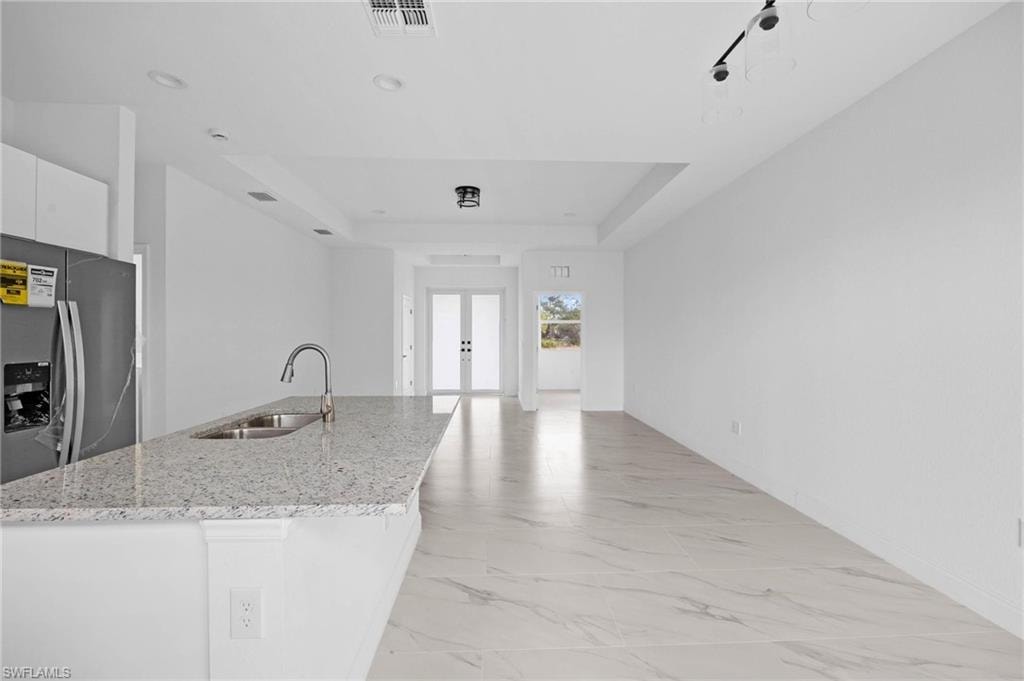Kitchen featuring stainless steel refrigerator with ice dispenser, a tray ceiling, white cabinetry, light stone countertops, and an island with sink