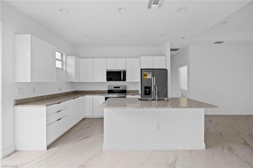 Kitchen with light stone countertops, modern cabinets, stainless steel appliances, a center island with sink, and white cabinetry