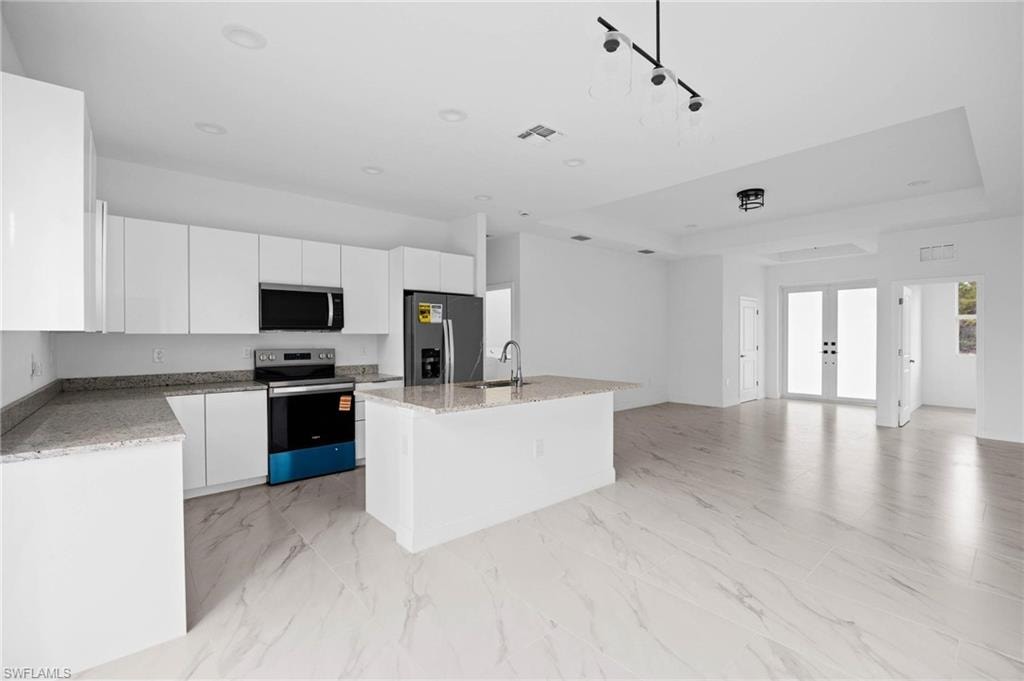 Kitchen with light stone countertops, stainless steel appliances, white cabinets, a kitchen island with sink, and a raised ceiling