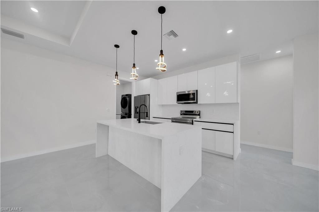 Kitchen featuring white cabinetry, stainless steel appliances, stacked washer and clothes dryer, a center island with sink, and light stone countertops