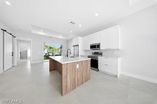 Dual tone kitchen featuring a barn door, two tone color scheme, a kitchen island with sink, stainless steel appliances, and a tray ceiling
