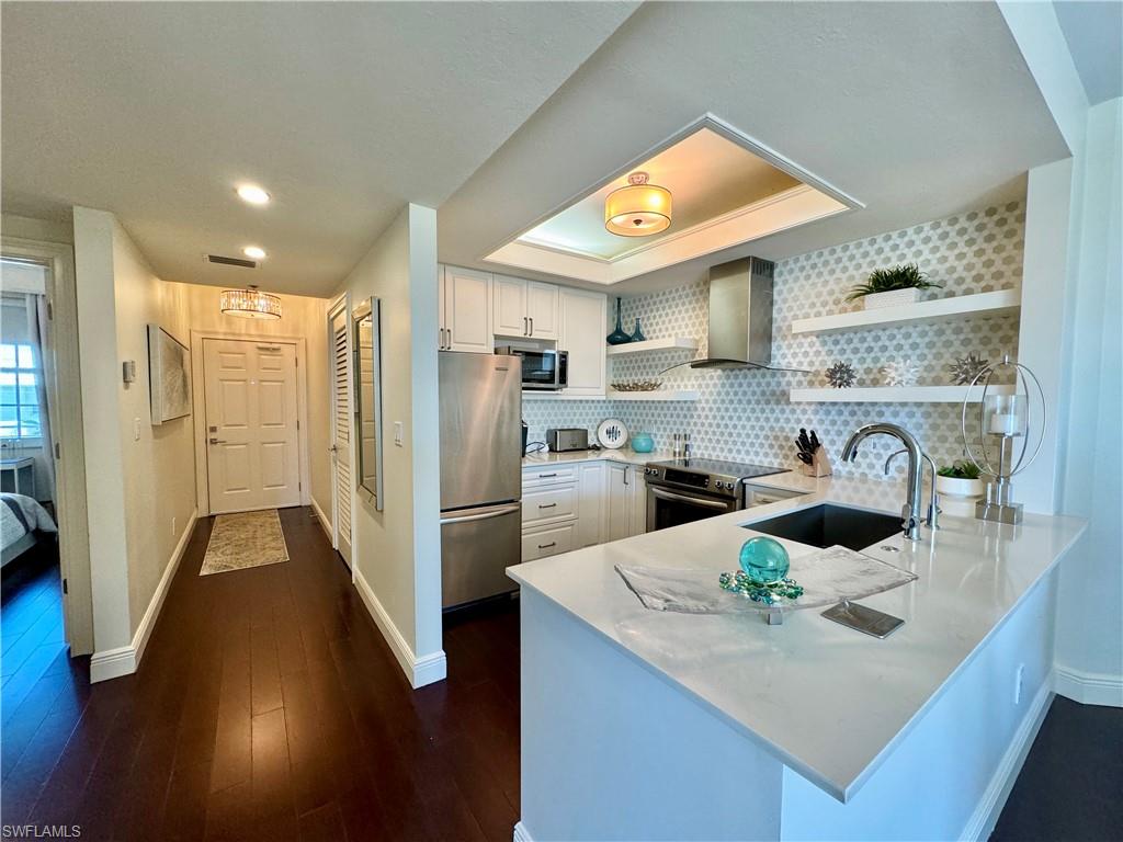 Kitchen with kitchen peninsula, stainless steel appliances, wall chimney exhaust hood, white cabinets, and dark wood-type flooring
