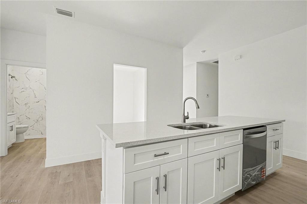 Kitchen with light wood-style floors, white cabinets, light stone countertops, an island with sink, and dishwasher