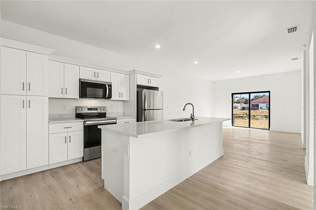 Kitchen featuring stainless steel appliances, white cabinetry, light wood-type flooring, an island with sink, and light stone counters