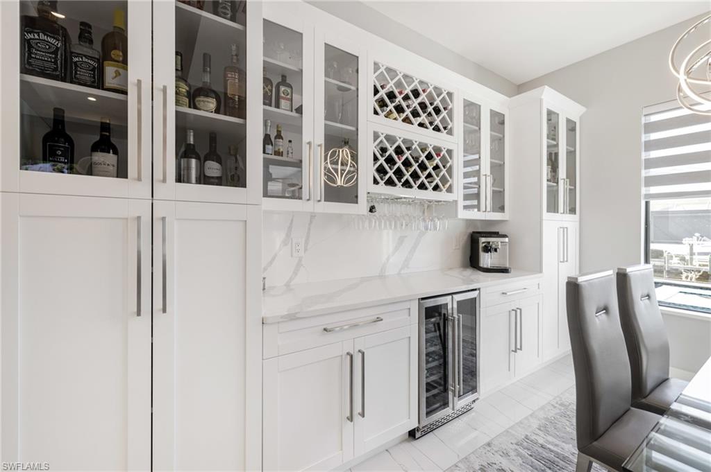 Indoor dry bar with beverage cooler, light stone countertops, white cabinets, glass fronted cabinets, and tasteful backsplash