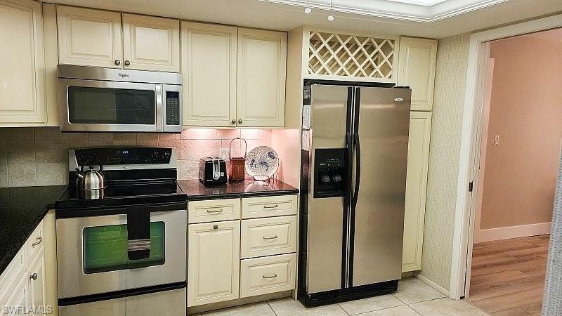 Kitchen with stainless steel appliances, backsplash, cream cabinetry, light tile patterned flooring, and dark stone countertops