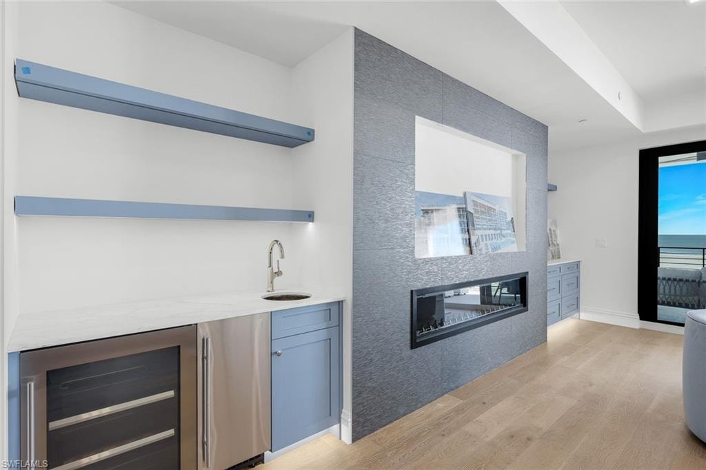 Indoor wet bar featuring open shelves, wine cooler, light stone counters, light wood finished floors, and stainless steel refrigerator