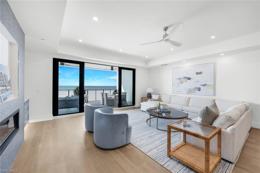 Living area featuring ceiling fan, a water view, light wood-style flooring, a fireplace, and a tray ceiling