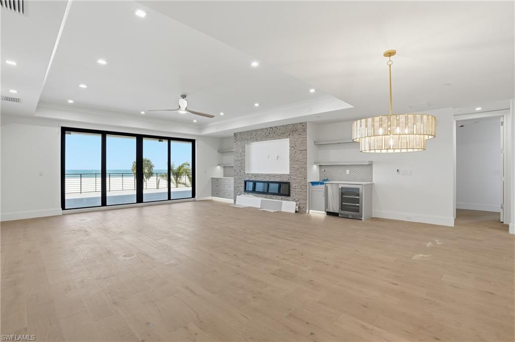 Unfurnished living room with light wood-type flooring, a stone fireplace, beverage cooler, a raised ceiling, and ceiling fan