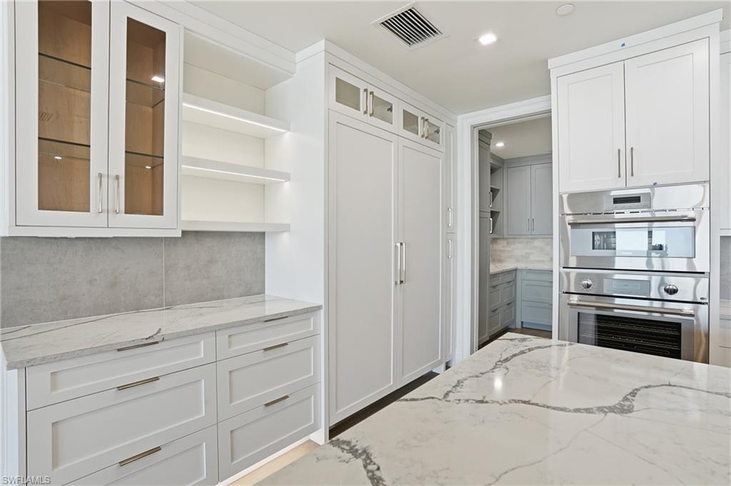 Kitchen with decorative backsplash, light stone countertops, open shelves, glass fronted cabinets, and stainless steel double oven