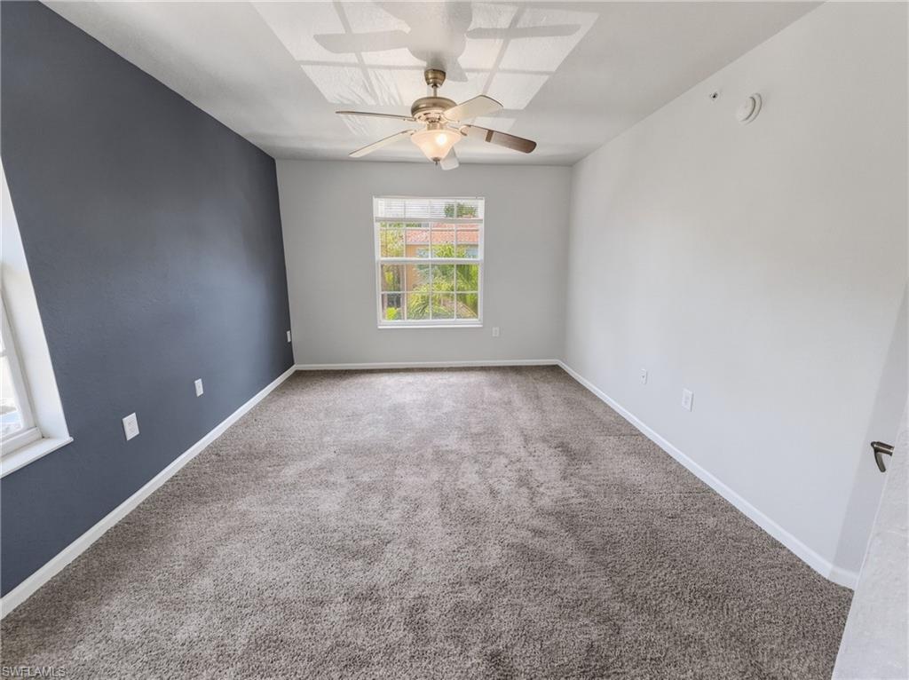Spacious room featuring plush gray carpeting, a window offering natural light, a ceiling fan with integrated lighting, and a distinct accent wall