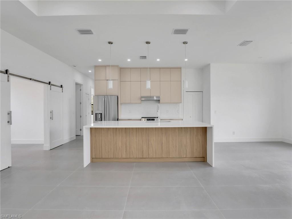 Kitchen with a barn door, a large island with sink, pendant lighting, light stone counters, and stainless steel refrigerator with ice dispenser