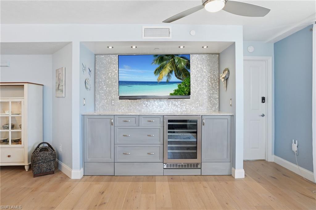 Bar with wine cooler, gray cabinets, light stone counters, light wood-style floors, and a ceiling fan - Virtually Edited Image