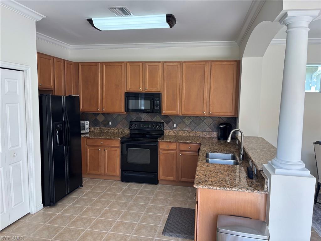 Kitchen with wood finish cabinetry, black appliances, ornate columns, dark stone counters, and a peninsula