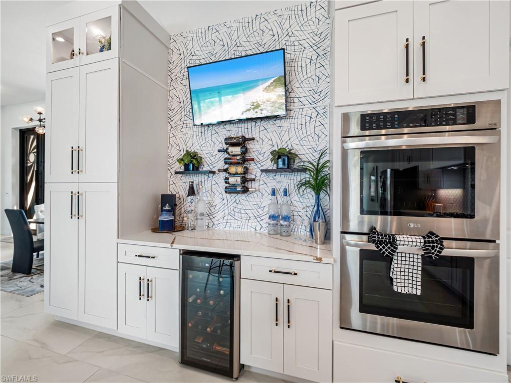 Bar with stainless steel double oven, wine cooler, white cabinetry, light stone counters, and glass fronted cabinets