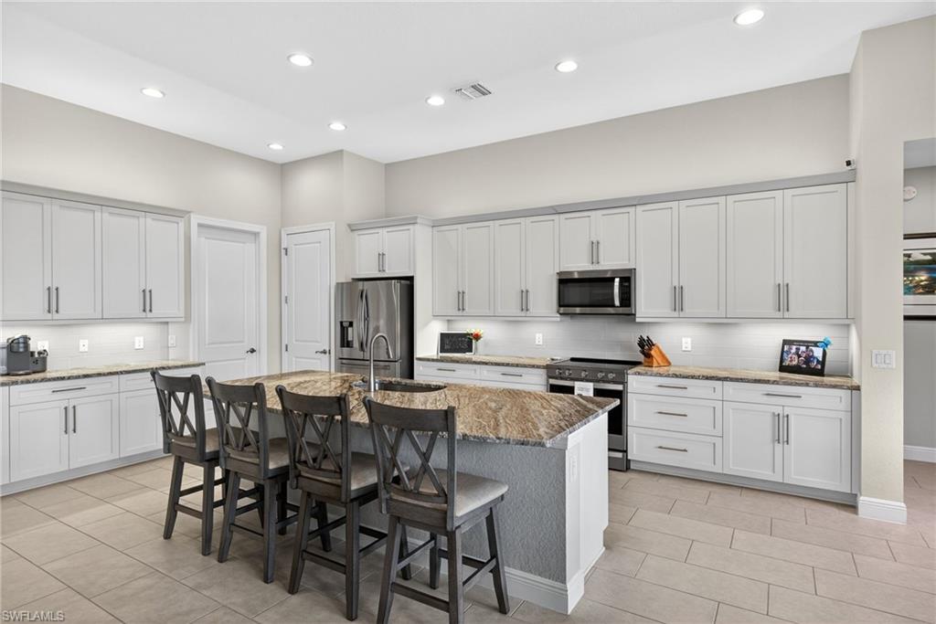 Kitchen with stainless steel appliances, a breakfast bar, light stone countertops, an island with sink, and white cabinets with under and upper cabinet lighting
