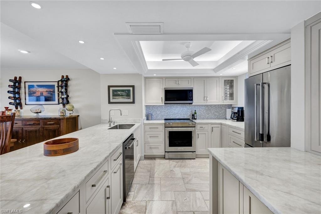 Kitchen with stainless steel appliances, a raised ceiling, light stone counters, a ceiling fan, and recessed lighting