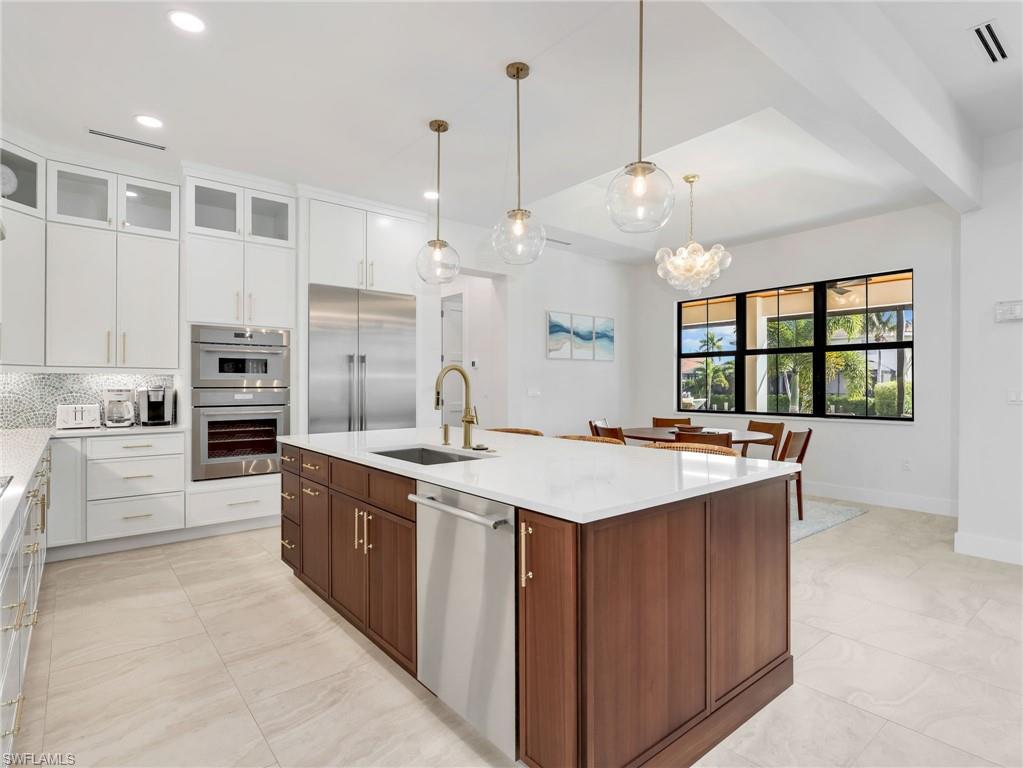 Two tone kitchen with two tone cabinets, glass insert cabinets, light stone counters, stainless steel appliances, and a center island with sink