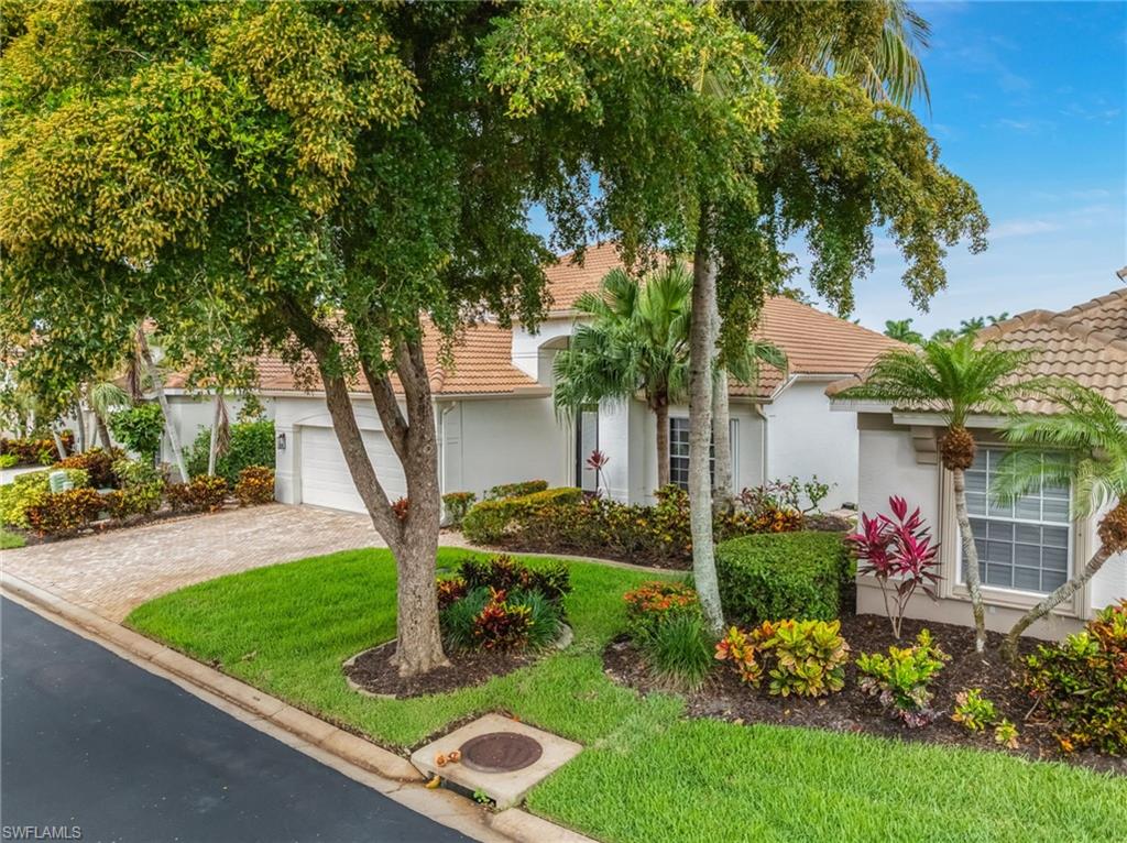Tile roof residence with a paver driveway, an integrated garage, and a lush front yard featuring mature trees, shrubs, and colorful plantings