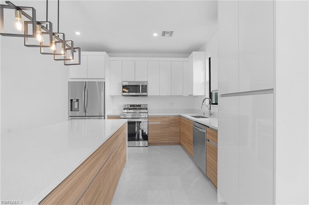 Two tone kitchen with stainless steel appliances, light stone counters, modern cabinets, decorative light fixtures, and light tile patterned flooring
