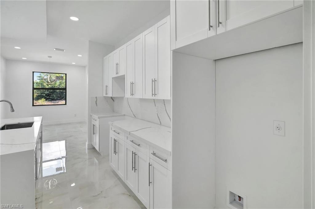 Kitchen featuring light stone counters, white cabinets, light marble finish floors, a center island with sink, and pendant lighting