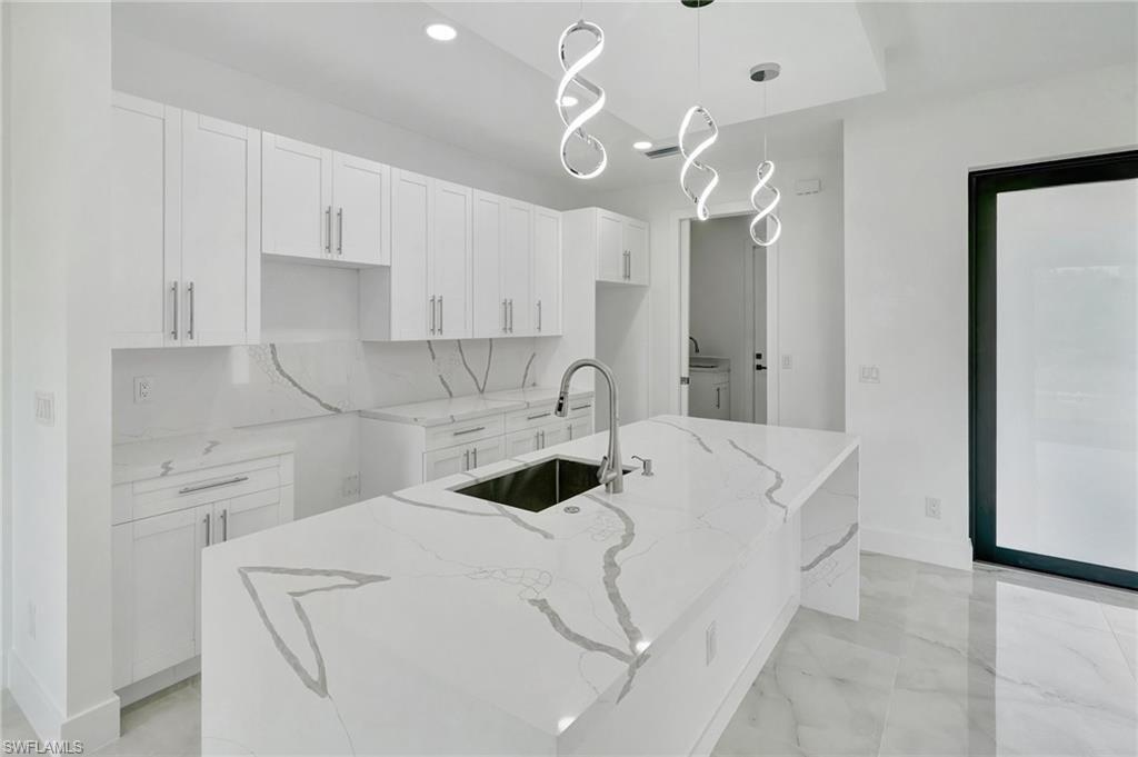 Kitchen featuring light stone counters, a center island with sink, white cabinetry, and decorative light fixtures