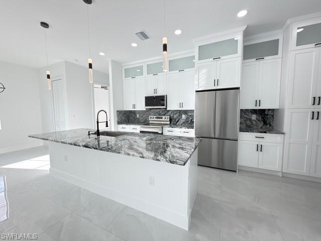 Kitchen with stainless steel appliances, glass fronted cabinets, dark stone counters, white cabinetry, and a large island with sink