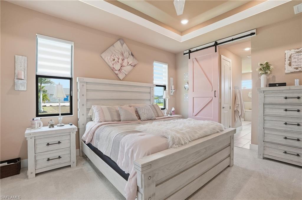 Bedroom with a barn door, multiple windows, light colored carpet, a tray ceiling, and a ceiling fan