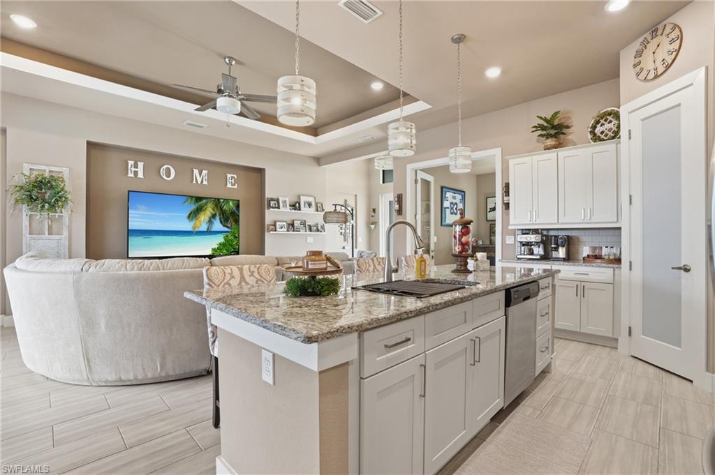 Kitchen with ceiling fan, open floor plan, light stone counters, an island with sink, and a raised ceiling