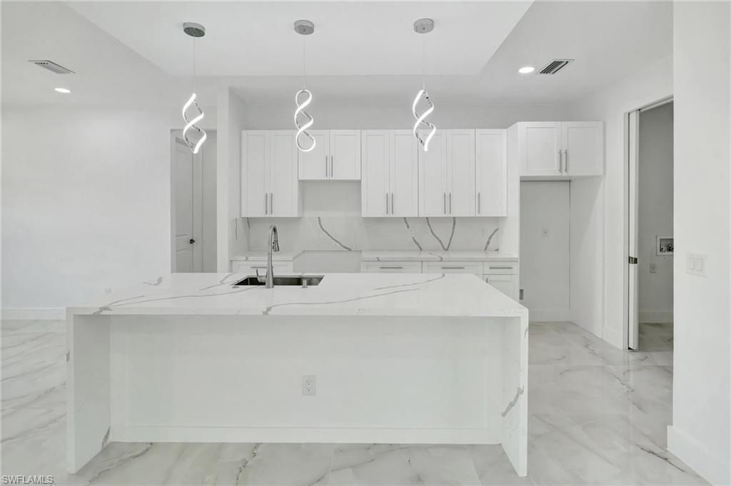Kitchen with light stone counters, a kitchen island with sink, decorative light fixtures, and white cabinets