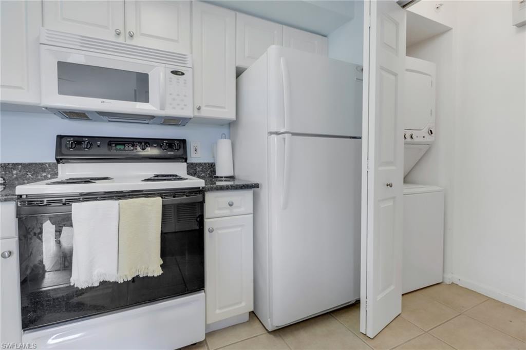 Kitchen with white appliances, white cabinetry, stacked washing machine and dryer, dark stone counters, and light tile patterned floors