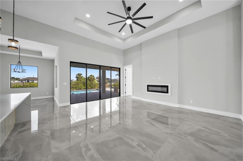 Unfurnished living room with a glass covered fireplace, a ceiling fan, recessed lighting, light marble finish flooring, and a high tray ceiling