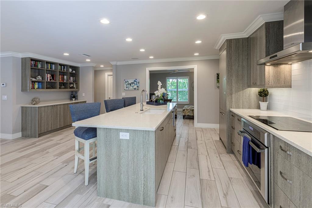 Kitchen with an island with sink, black electric cooktop, wall chimney exhaust hood, oven, and sink