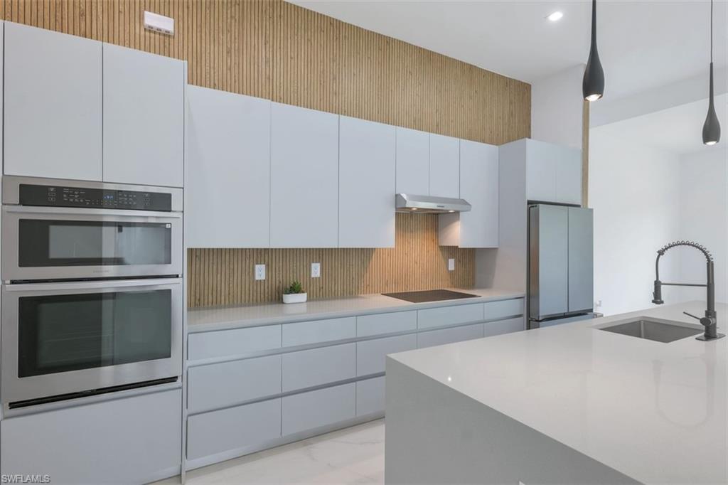 Modern kitchen featuring light gray cabinetry, stainless steel appliances, a wood-slat backsplash, a large island with an undermount sink, and contemporary pendant lighting