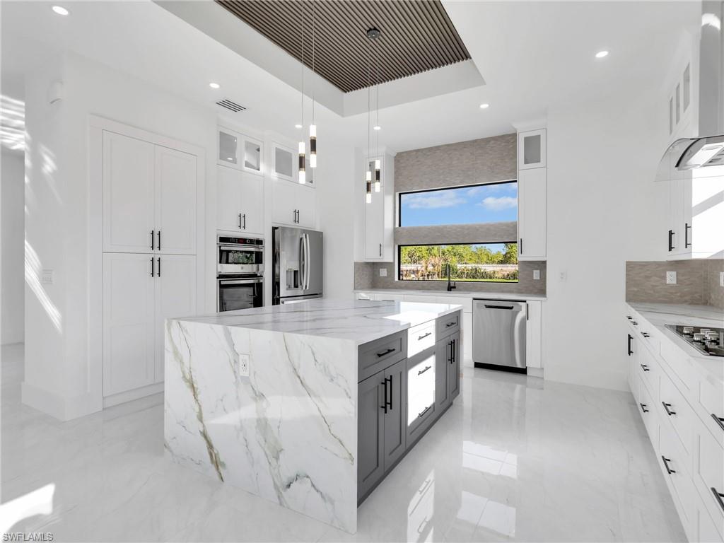 Dual tone kitchen with glass fronted cabinets, light stone counters, pendant lighting, a center island, and a raised ceiling