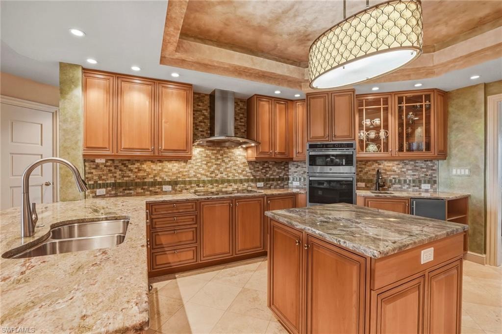 Kitchen with wood finish cabinetry, light stone counters, decorative light fixtures, stainless steel double oven, and a raised ceiling
