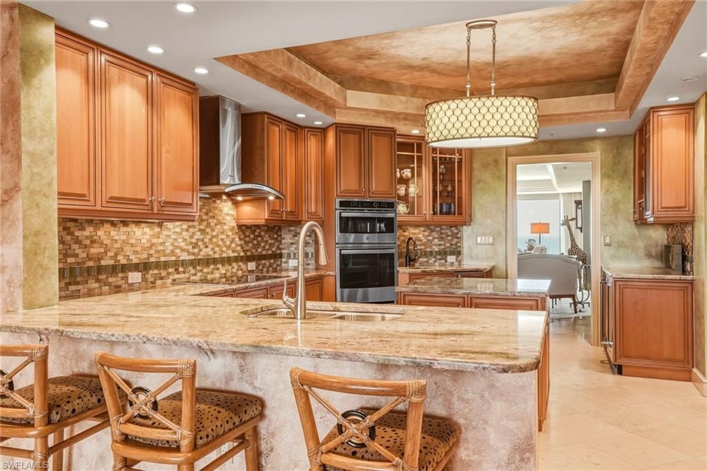Kitchen featuring decorative backsplash, light stone countertops, wood finish cabinets, a peninsula, and a raised ceiling