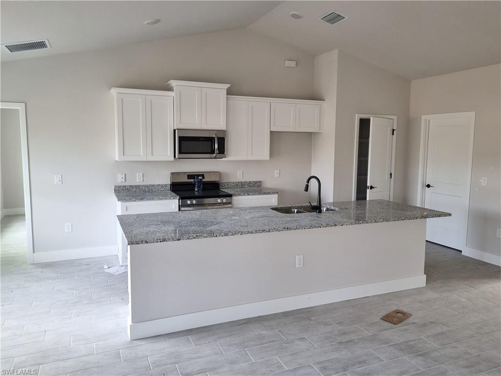 Kitchen featuring stainless steel appliances, light stone counters, white cabinetry, an island with sink, and lofted ceiling