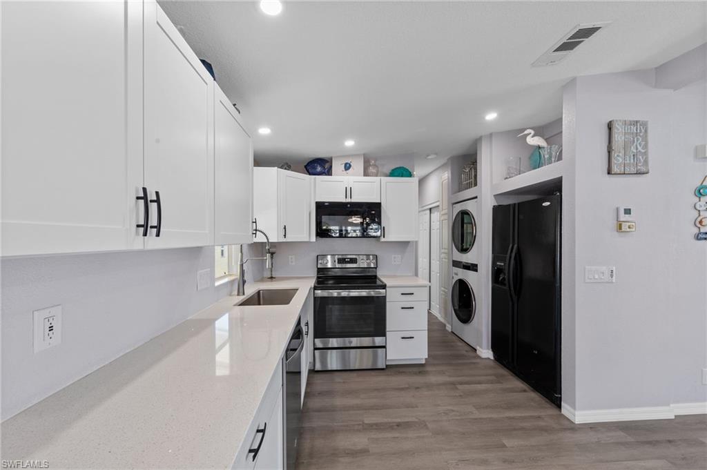 Kitchen with black appliances, stacked washer / drying machine, white cabinets, light stone counters, and light wood-style flooring